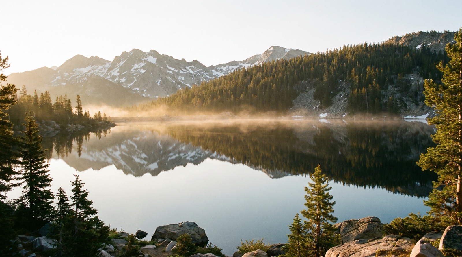 Wide-angle photographic view of a still mountain reservoir at golden hour with a forested ridge and high peaks reflected in the water, no logos, no signage, no people
