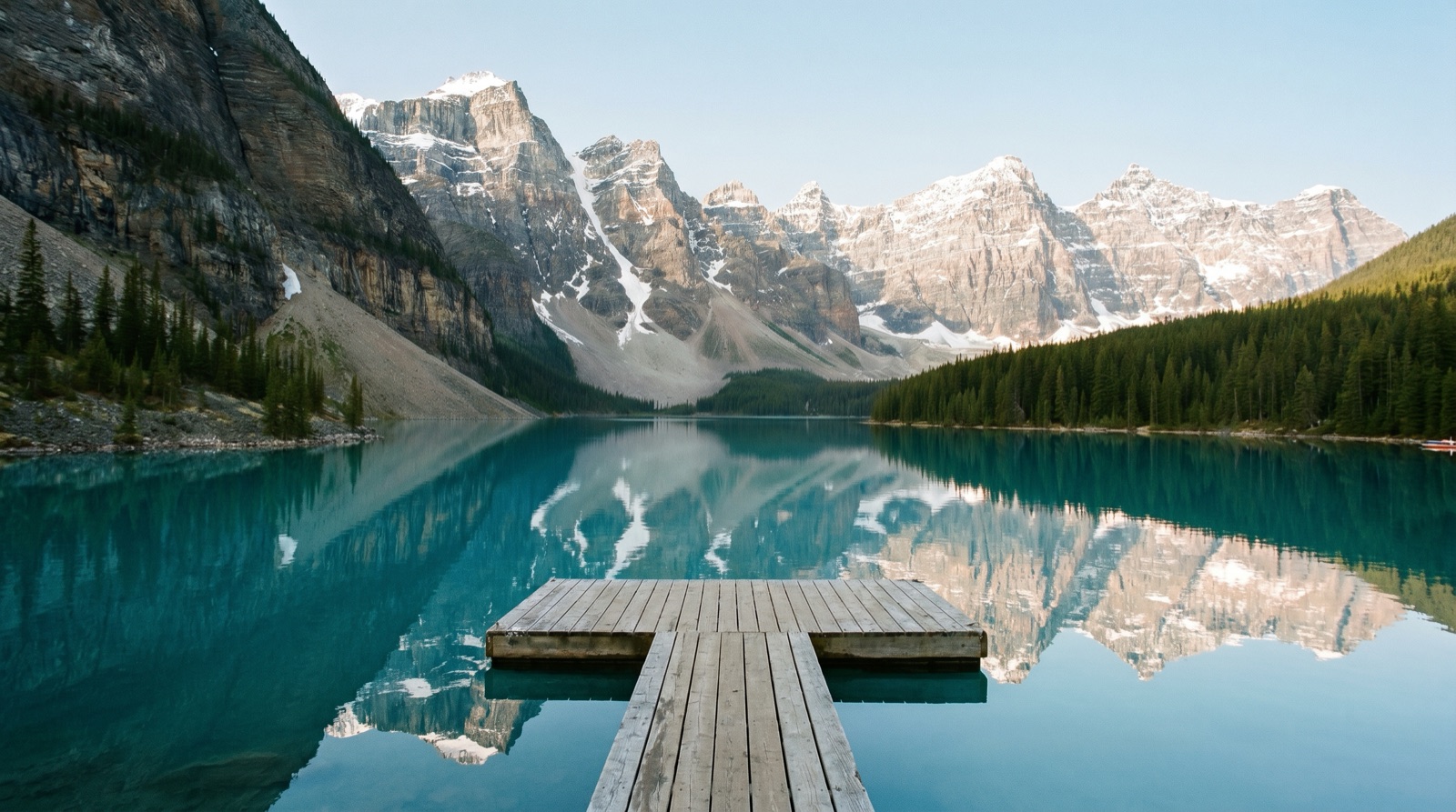 Wide photographic view of a turquoise glacial lake ringed by steep forested mountains with a small wooden dock in the foreground, no logos, no signage, no people