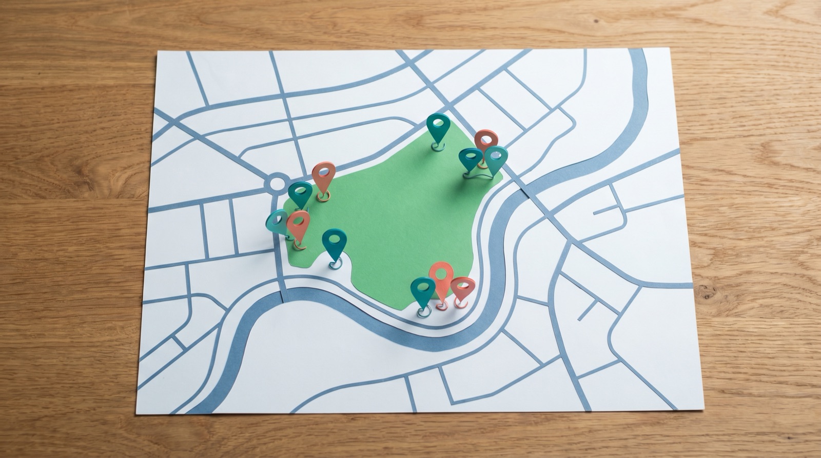 Photographic overhead view of a paper map-style city layout with several colored location pins clustered around a parkland area, illustrative, no real place names, no logos