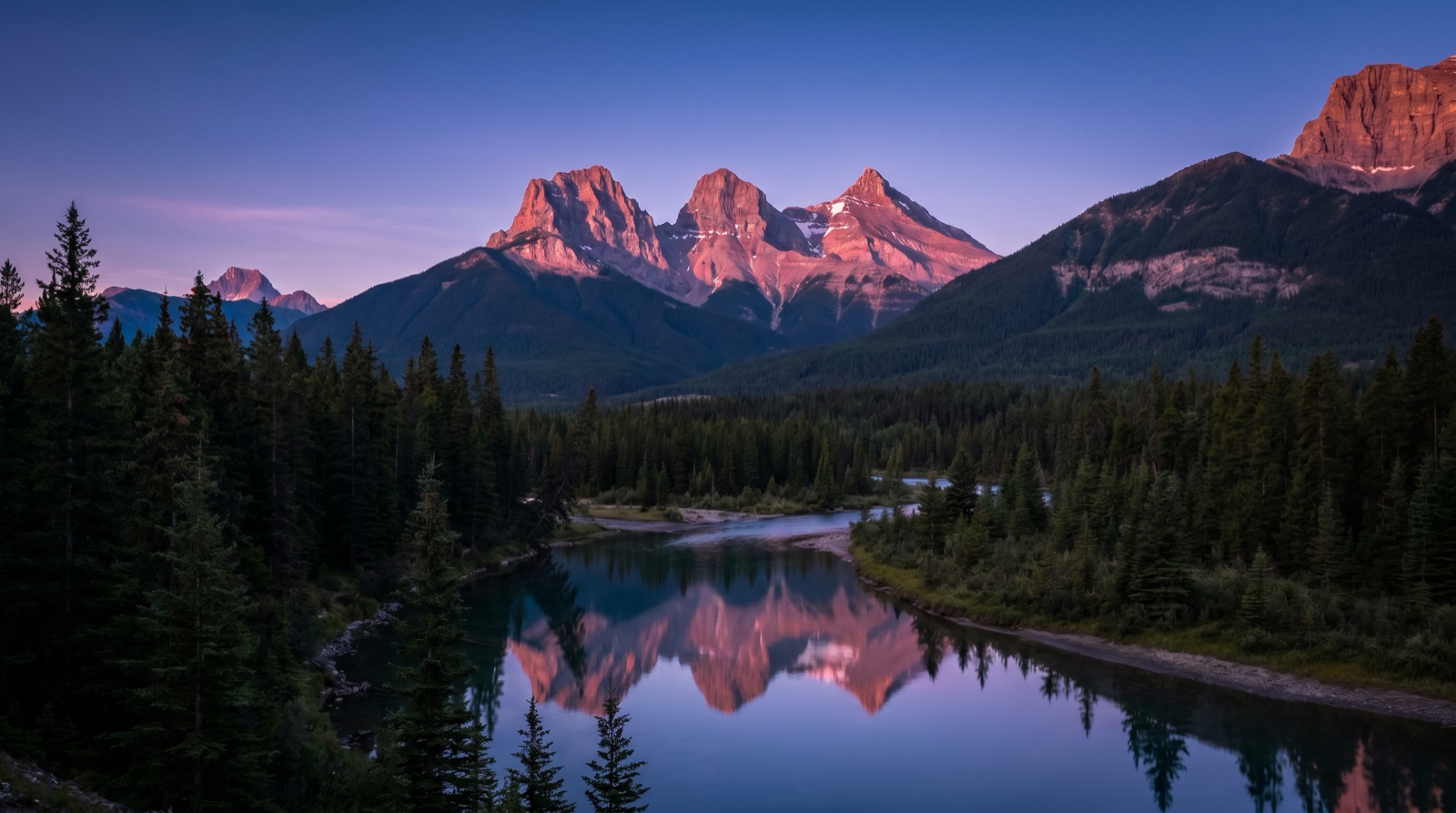 Wide-angle photographic view of three jagged mountain peaks at dusk above a pine valley with a calm river in the foreground, no logos, no signage, no people