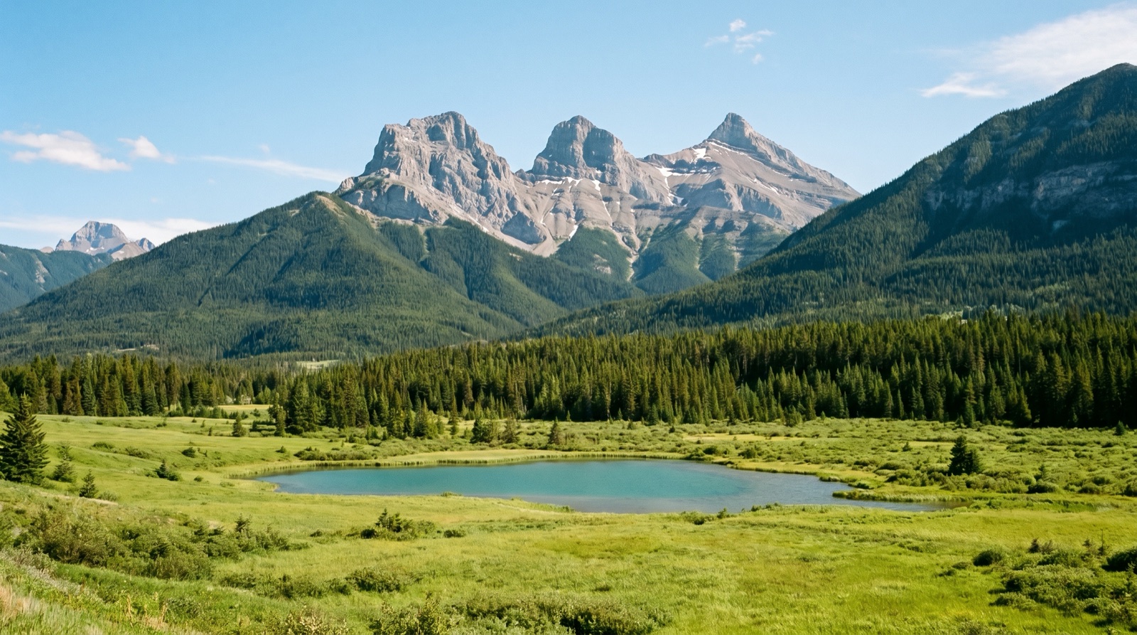 Wide photographic view of a broad green mountain valley in summer with a turquoise lake and three steep rocky peaks rising behind a pine forest, no logos, no signage, no people