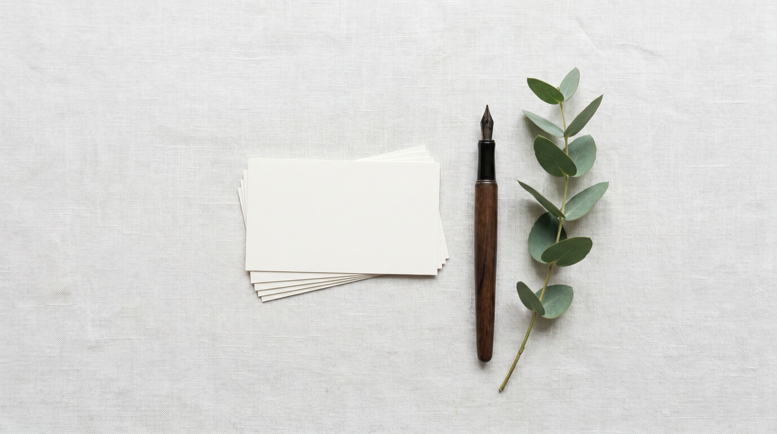 Photographic overhead of a stack of blank cream place cards, a fountain pen, and a sprig of eucalyptus on a linen surface, soft daylight, no logos, no text, no people