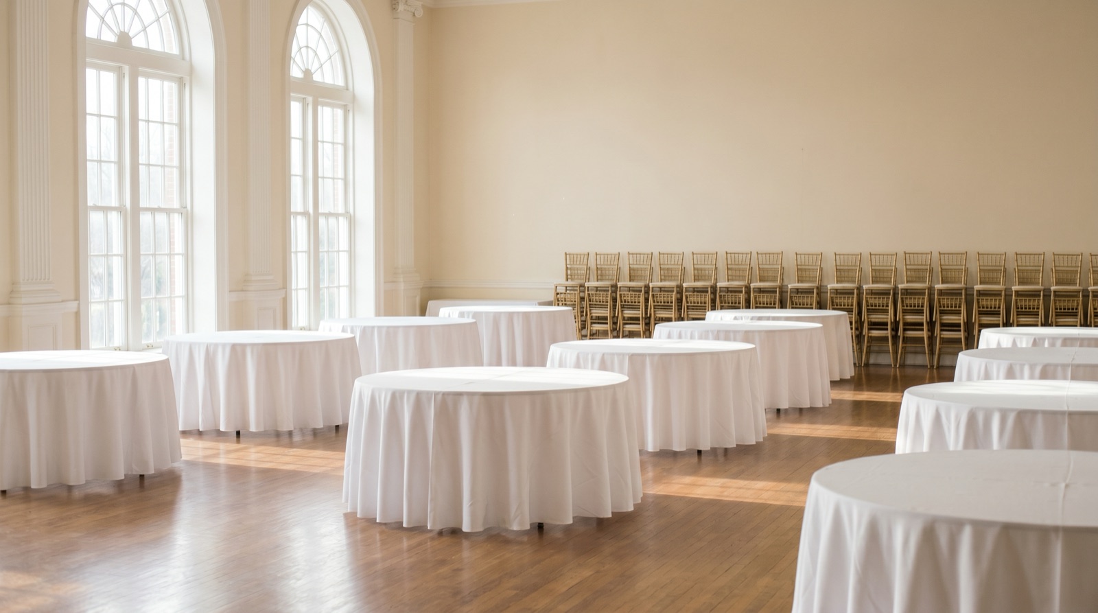 An empty elegant reception hall before setup, with bare round tables draped in white linen on a wood floor, stacked banquet chairs along the wall, and soft morning light through tall arched windows