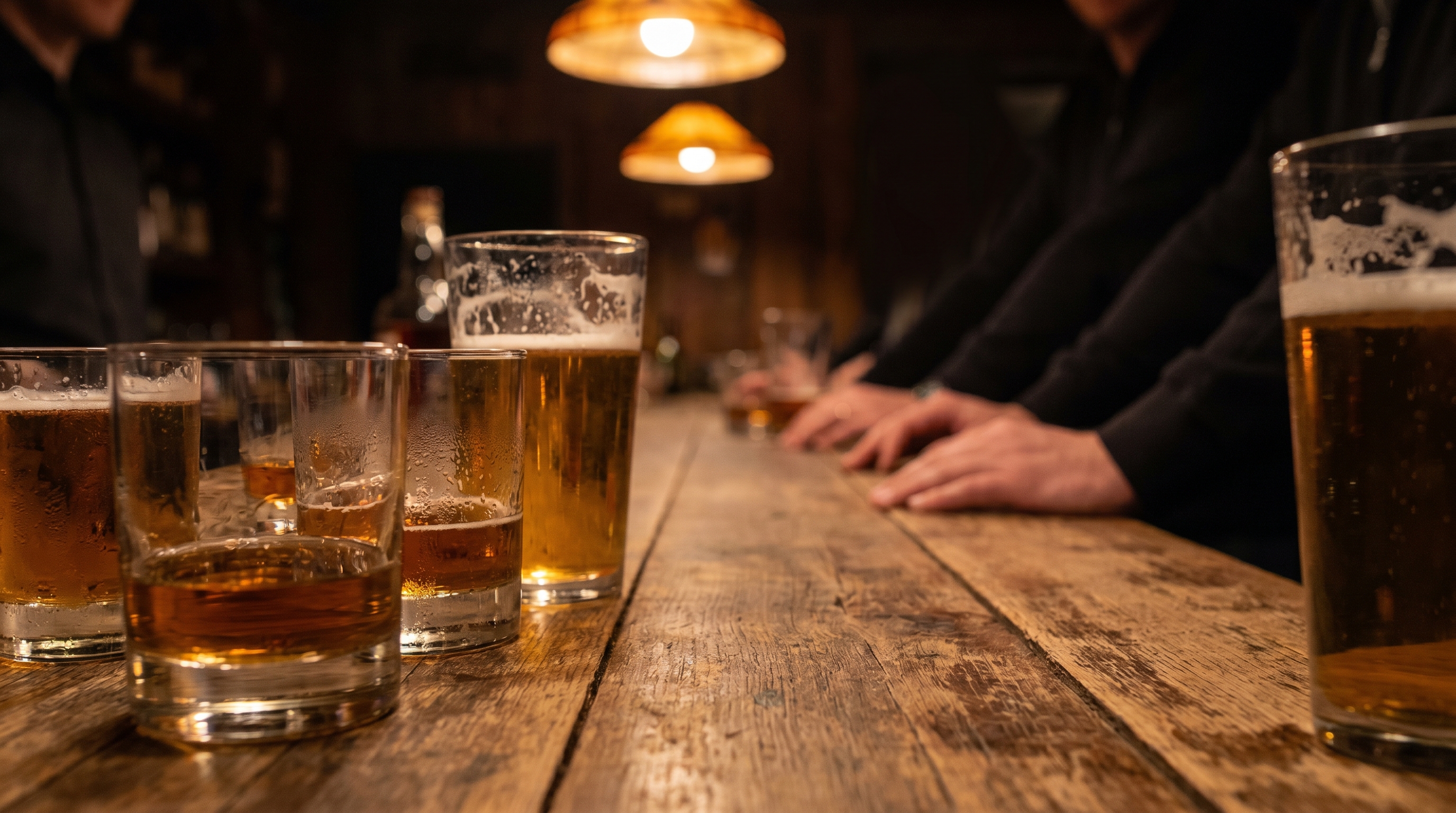 Wooden bar in warm low light with several glasses of bourbon and beer, anonymous hands resting on the bar in soft focus