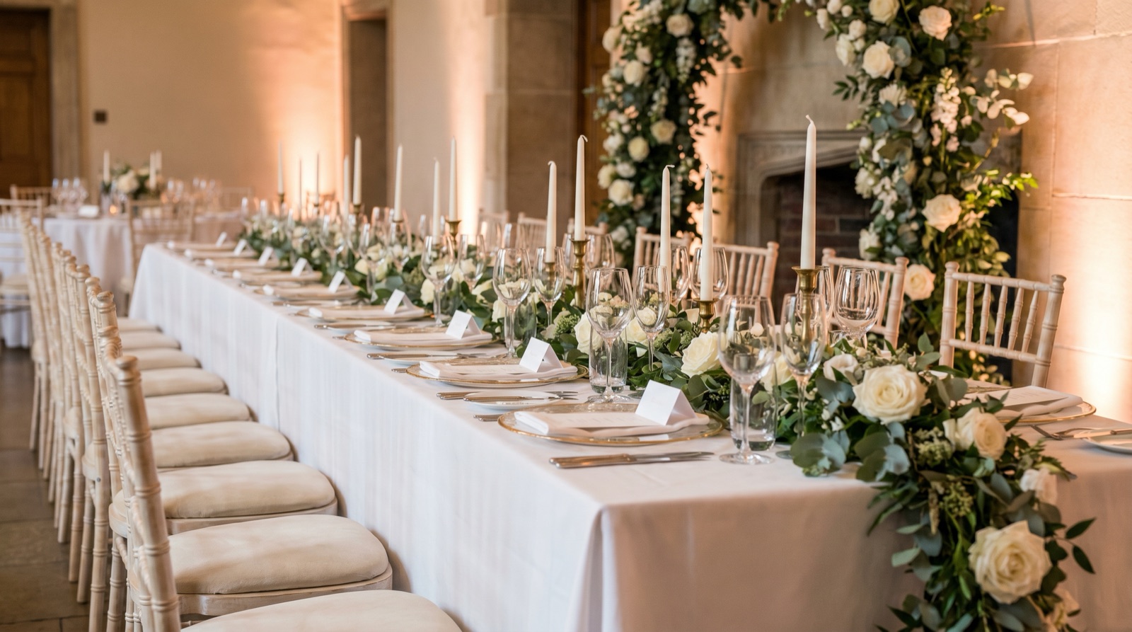 An elegant wedding head table draped in white linen with cushioned chairs, gold chargers, crystal stemware, tall taper candles, a lush eucalyptus-and-white-rose garland, and small blank place cards under soft romantic lighting