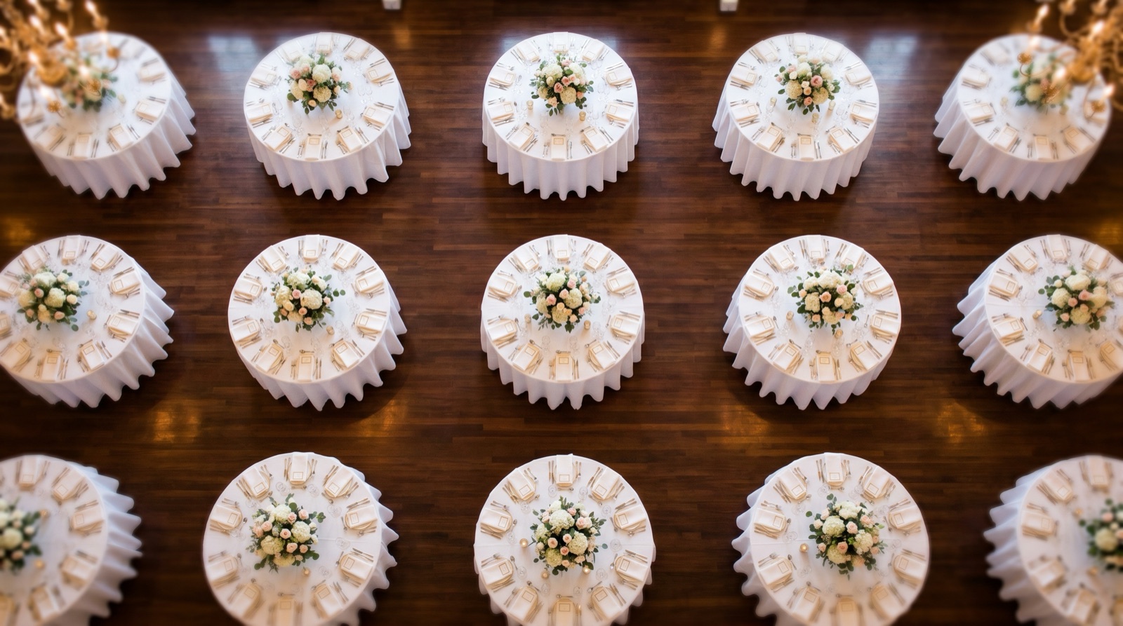 An overhead view of round wedding reception tables arranged in a neat symmetrical grid across a polished wood ballroom floor, each set identically with white linens, blush-and-white floral centerpieces, gold-rimmed plates, and place cards